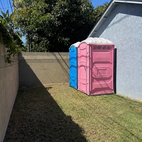 A blue and a pink porta potty placed on a backyard lawn along a fence