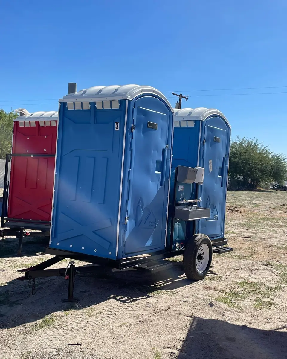 Blue porta potties set beside a red utility trailer on a sunny jobsite