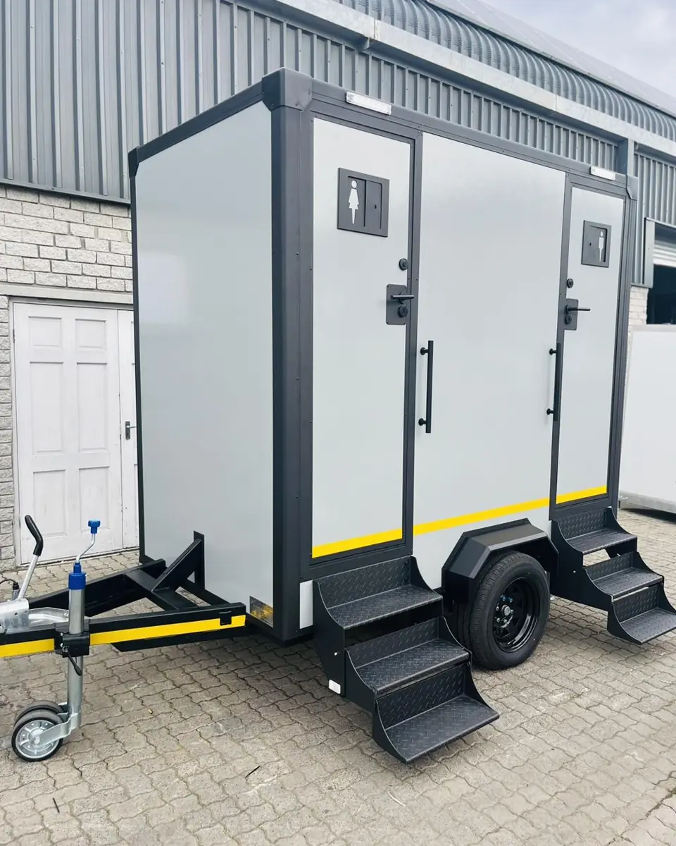 Gray two-station restroom trailer with fold-down steps parked on pavers