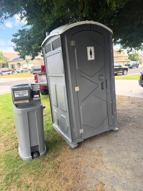 Gray standard porta potty next to a freestanding hand wash station on a lawn