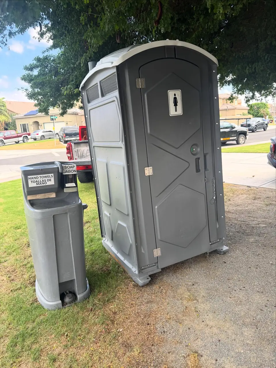 Gray standard porta potty next to a freestanding hand wash station on a lawn